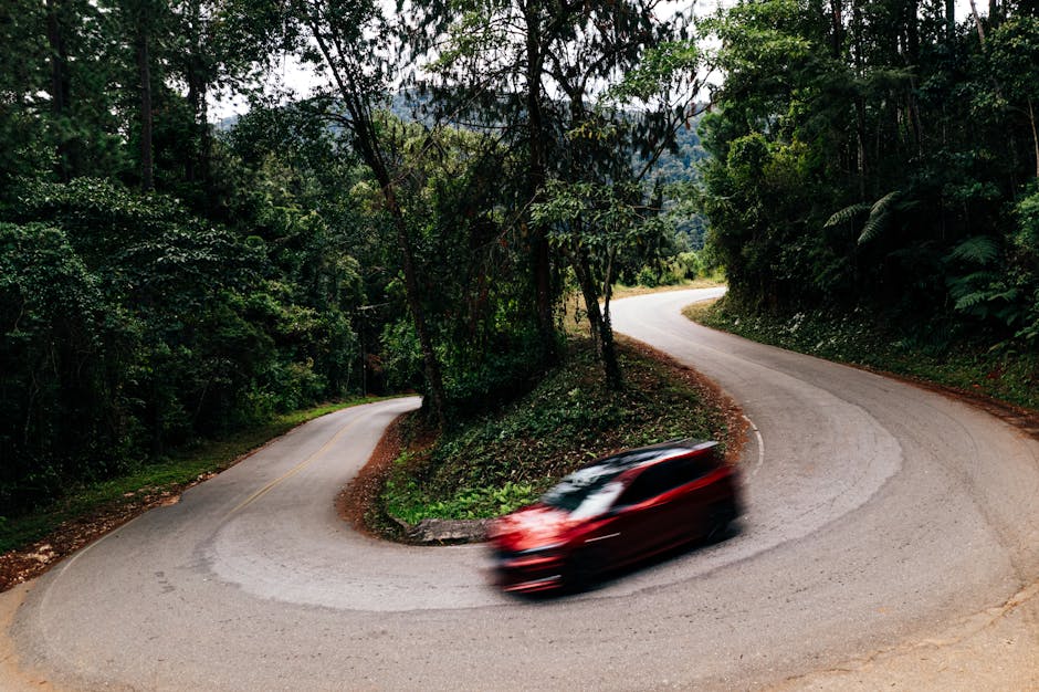 An aerial view showing a narrow road with three white vehicles parked along the curb, bordered by dense greenery and rocky terrain on either side. The road appears to be within a residential or semi-r