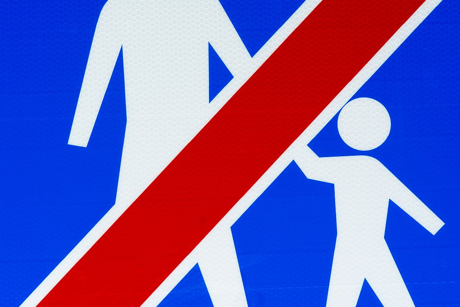 Close-up of a blue and white pedestrian crossing sign with a red diagonal line crossing out two figures of walking people, indicating no pedestrian access. The sign is mounted outdoors on a surface, w