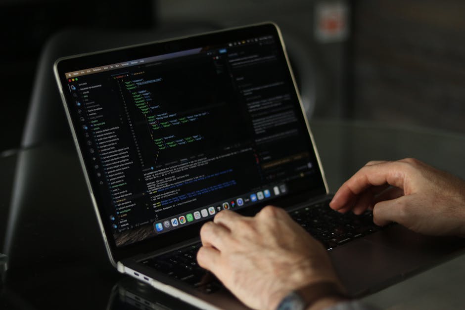 A close-up of a person's hand resting on the keyboard of a silver laptop placed on a wooden surface, with the screen displaying lines of code in a text editor. The person is wearing a black wristwatch