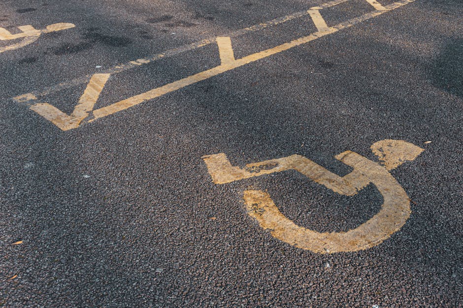 The image depicts a section of an outdoor parking lot with a visible yellow marked accessible parking space, including a wheelchair symbol painted on the dark asphalt surface. Surrounding the marked a